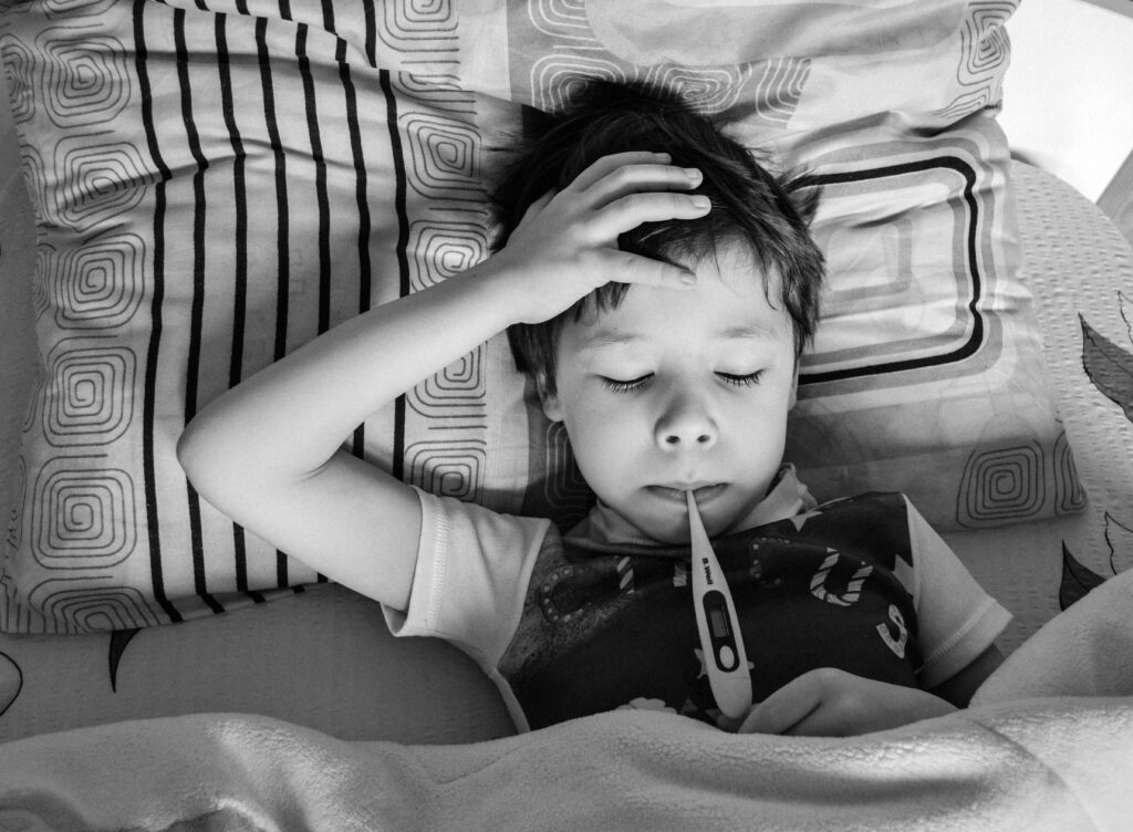 Black and white overhead view of child in casual clothing measuring body temperature with electronic thermometer while lying on bed under blanket